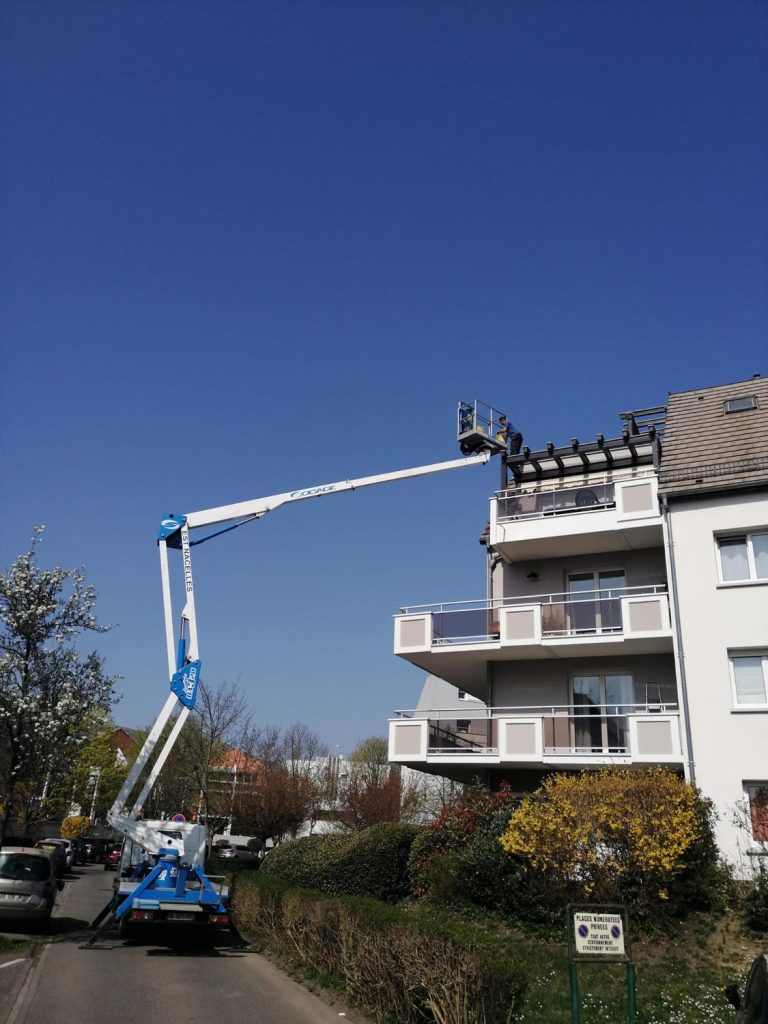 répare toiture - Un ouvrier dans une nacelle bleue inspecte ou répare le toit d'un immeuble résidentiel à plusieurs étages par une journée claire et ensoleillée.