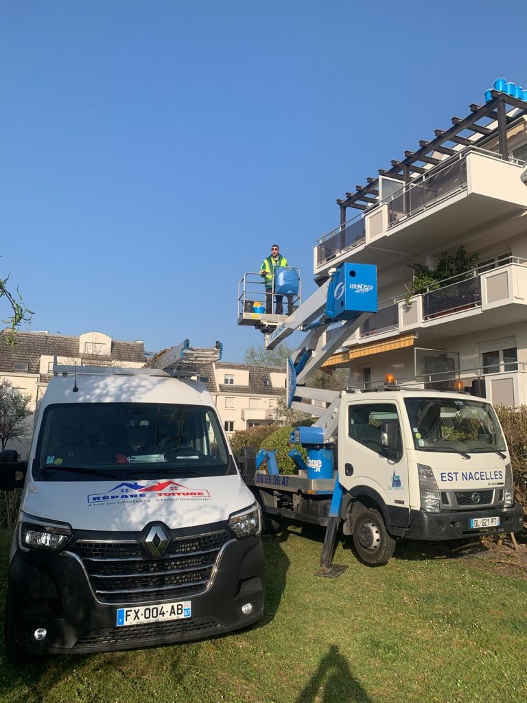 répare toiture - Un travailleur portant un gilet de sécurité utilise une nacelle élévatrice bleue près d'un immeuble résidentiel. Deux véhicules de service sont garés sur l'herbe à proximité.