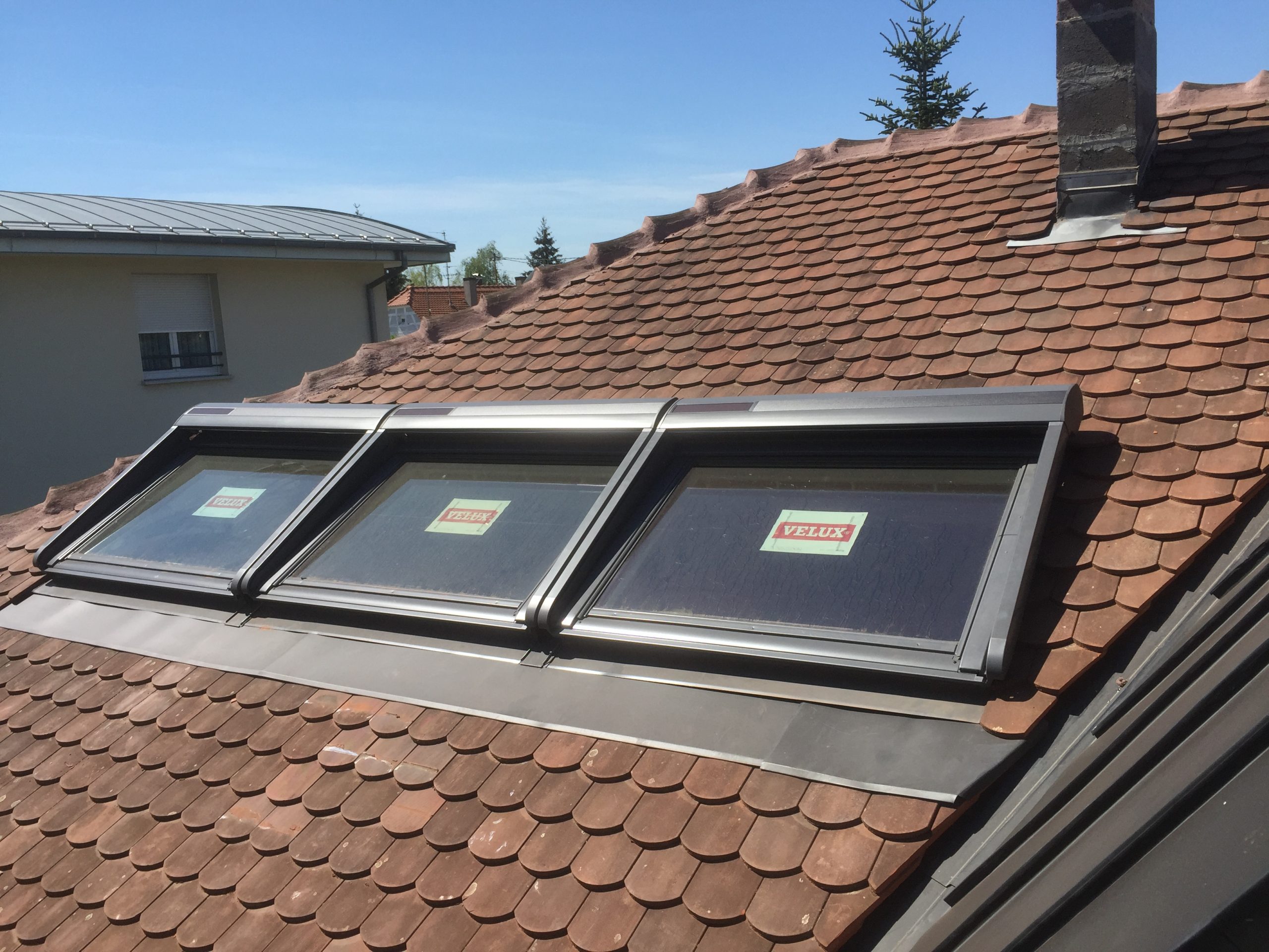 répare toiture - Trois fenêtres de toit Velux installées côte à côte sur un toit en pente avec des bardeaux bruns, sous un ciel bleu clair.