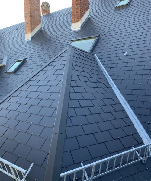répare toiture - Un toit d'ardoise gris foncé avec plusieurs cheminées en briques, des fenêtres triangulaires et des pare-neige en métal près du bord.